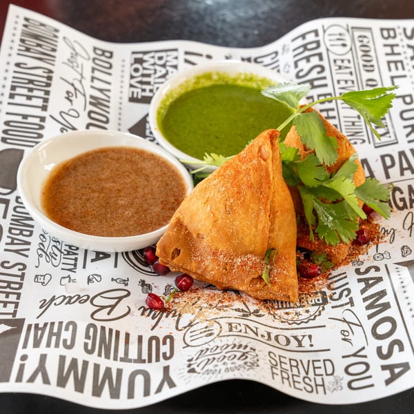 Two golden-brown samosas with cilantro, pomegranate seeds, and two dipping sauces on branded paper.