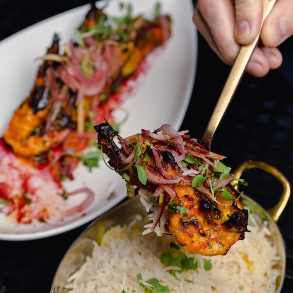 A golden spoon lifts a piece of grilled Indian chicken with red onions and herbs over rice.