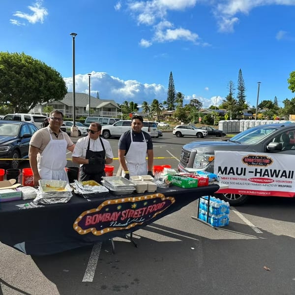 Three Bombay Street Food volunteers stand behind a table with food and drinks for wildfire relief in Maui.