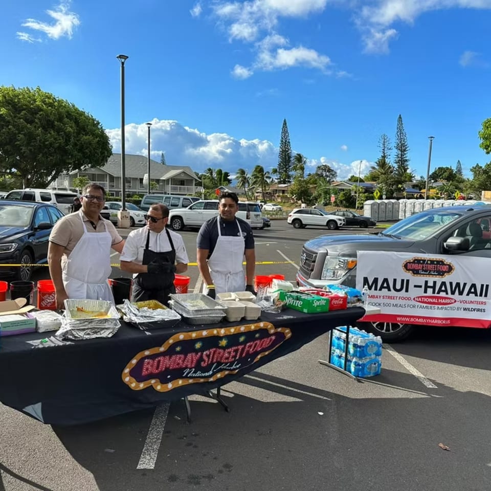 Three Bombay Street Food volunteers stand behind a table with food and drinks for wildfire relief in Maui.