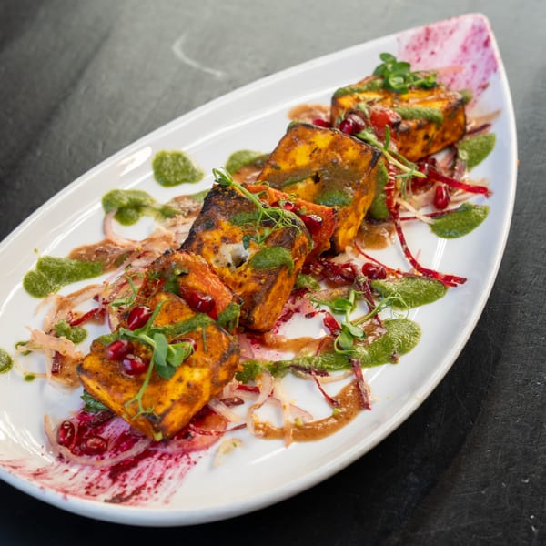 Indian paneer tikka, garnished with green sauce, pomegranate seeds, and microgreens on a white plate.