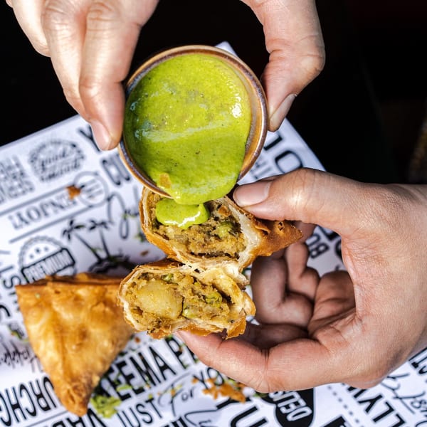 Hands pouring green chutney over a broken samosa, revealing its potato and pea filling.