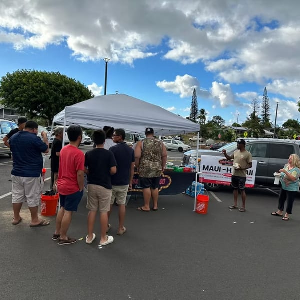 People gathered around a white tent and a truck with a "Maui-H" banner, under a cloudy sky.