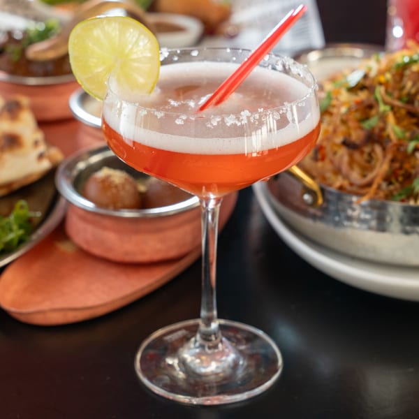 A red cocktail with a lime slice and red straw, surrounded by Indian dishes at Bombay Street Food.