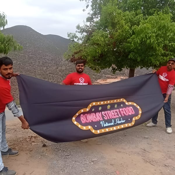 Three men in red shirts hold a "Bombay Street Food National Harbor" banner outdoors.