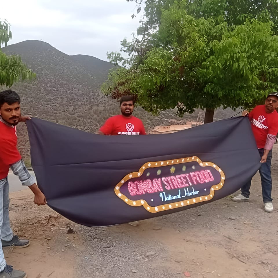 Three men in red shirts hold a "Bombay Street Food National Harbor" banner outdoors.