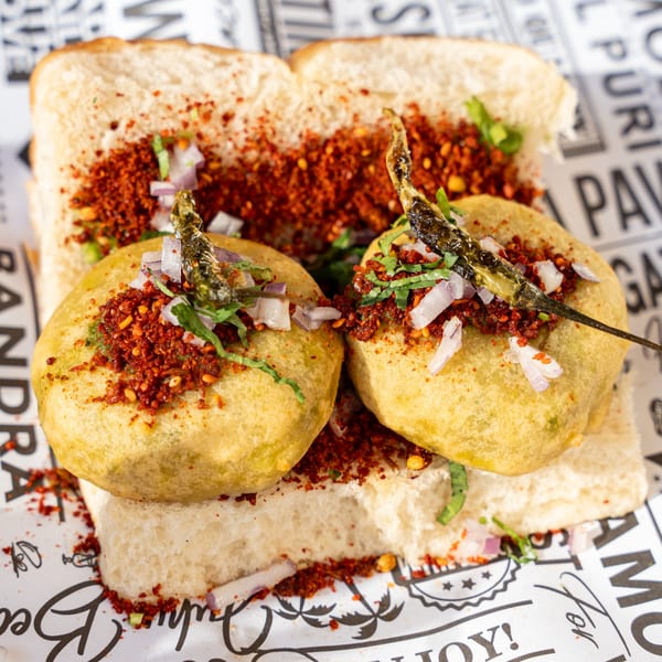 Two Bombay Vada Pavs, fried potato fritters in bread, topped with spices, onions, and green chilies.