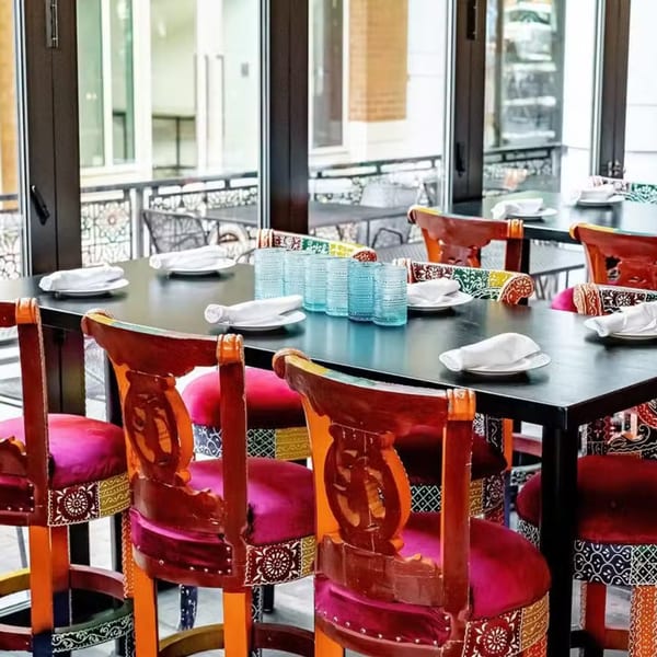 Bombay Street Food's dining area with colorful chairs, dark tables, and blue glasses, next to large windows.