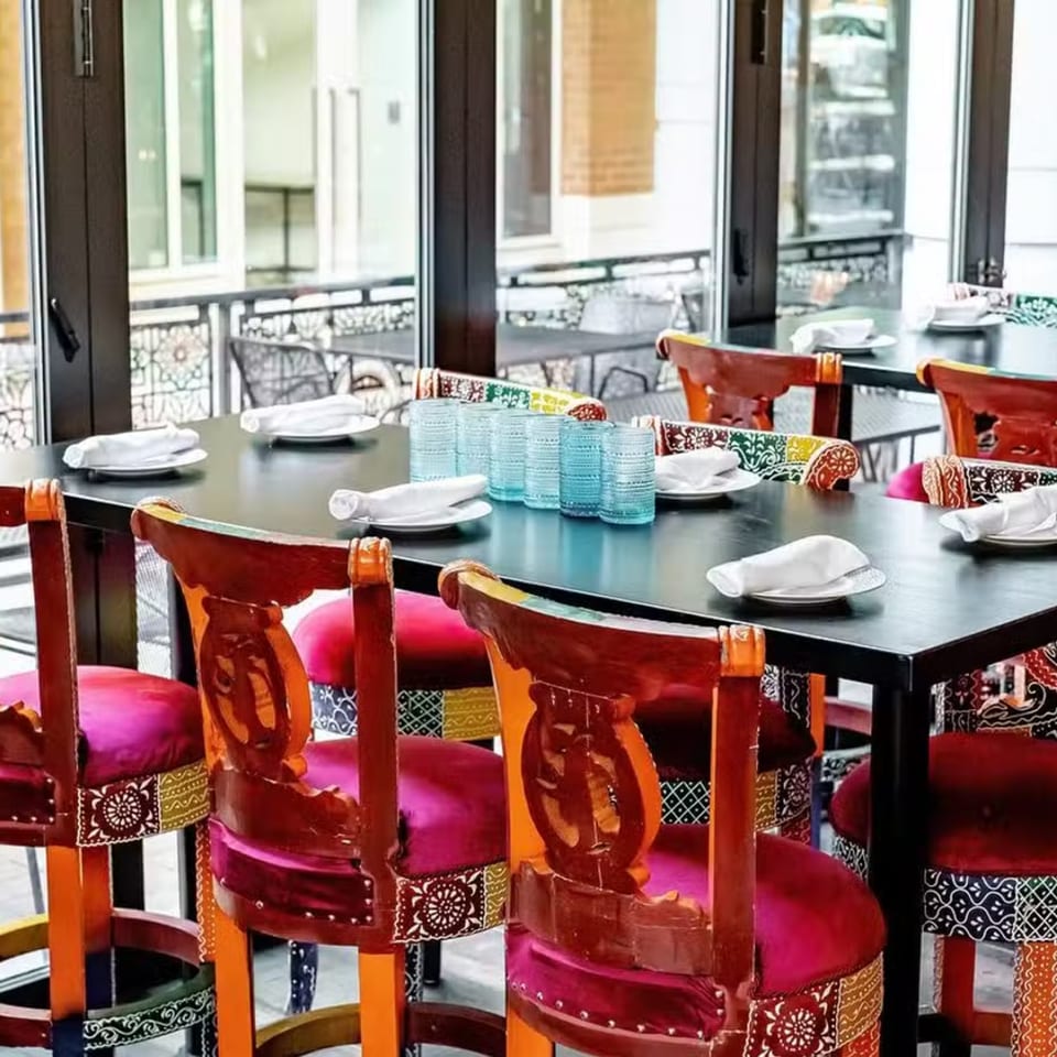 Bombay Street Food's dining area with colorful chairs, dark tables, and blue glasses, next to large windows.